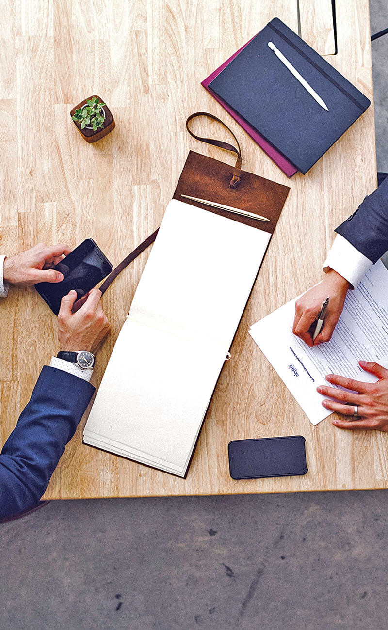 Professionele samenwerking binnen een gestructureerde aanpak Bovenaanzicht van een professionele vergadertafel met documenten, notitieblokken, smartphone en twee personen die samenwerken. De afbeelding ondersteunt het idee van een gestructureerde aanpak, analyse en duidelijke toepassing binnen een professionele omgeving.